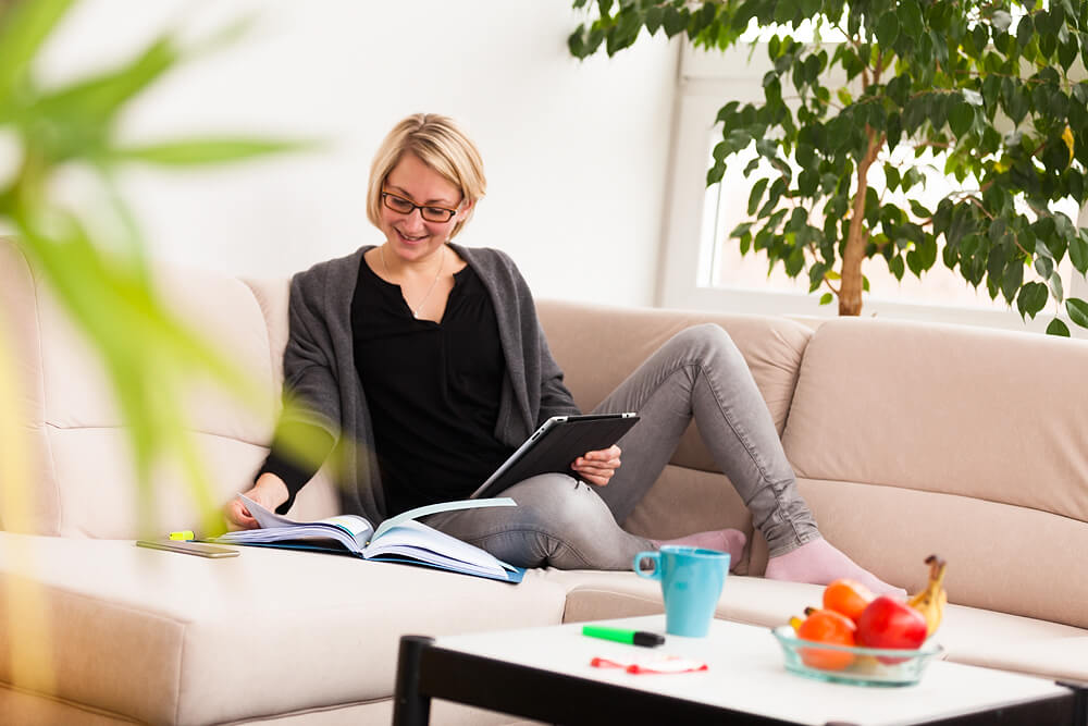 Ein Studierende, die auf einem Sofa sitzt und in einem Buch liest. In der Hand hält Sie ein Tablett.