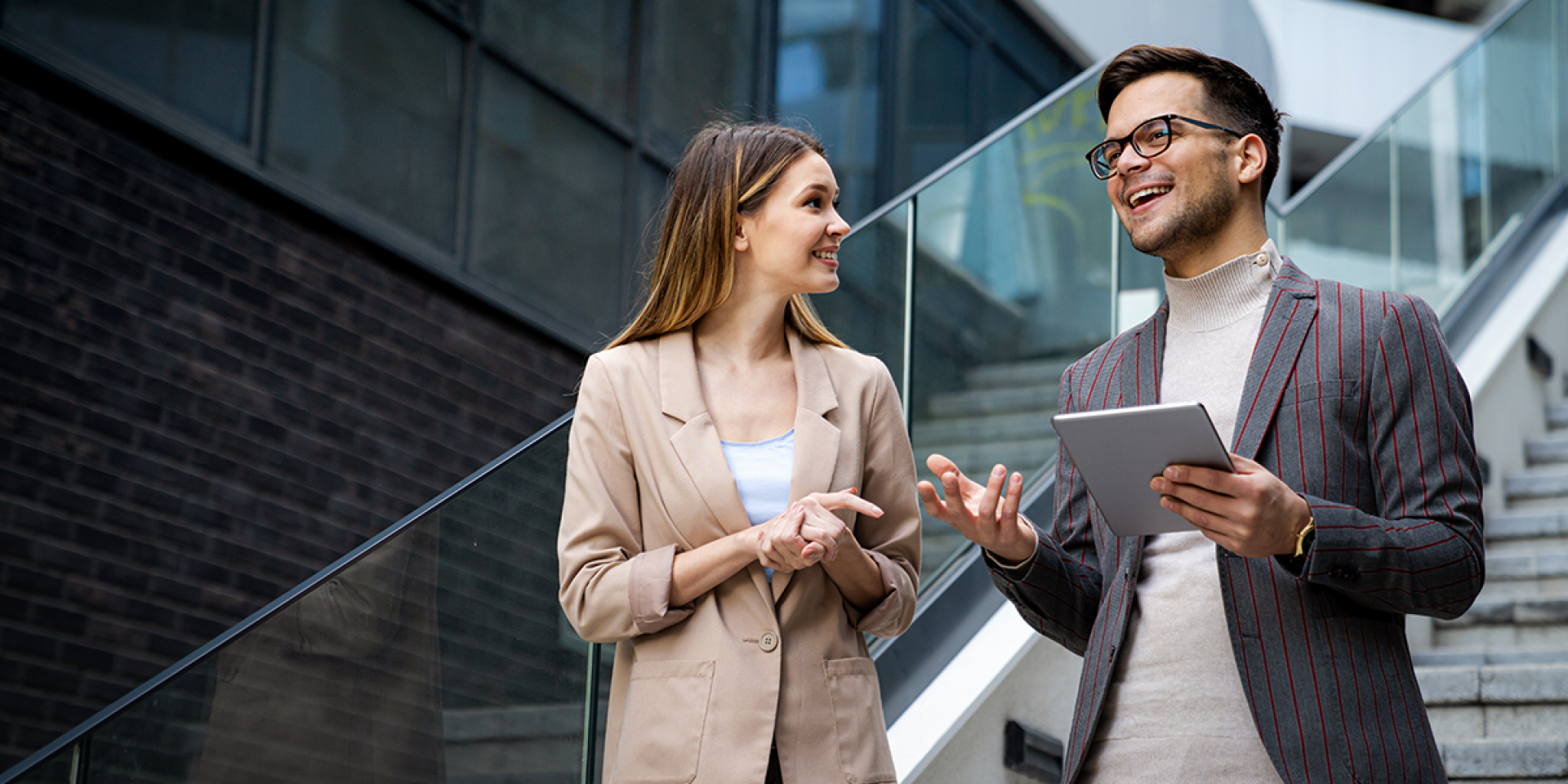 Eine Frau und ein Mann mit Tablet, beide im Business Look, stehen auf einer Treppe in einem modernen Unternehmen und unterhalten sich
