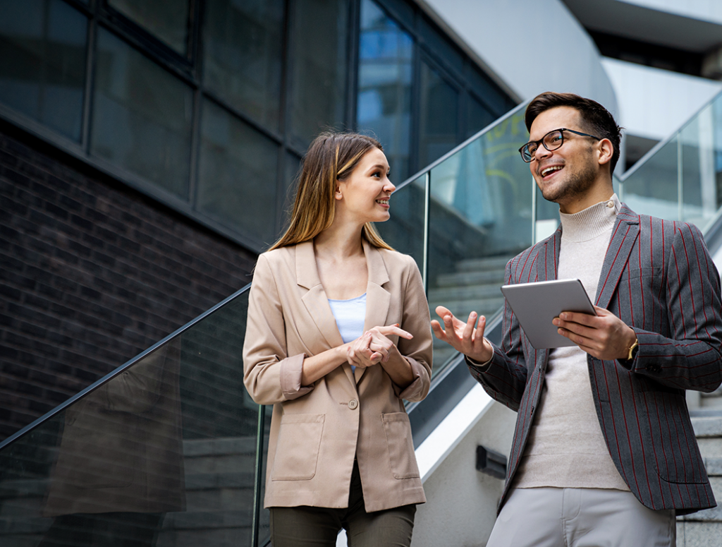 Master Wirtschaftsrecht (Bau und Immobilien) Eine Frau und ein Mann mit Tablet in der Hand, beide im Business Look, stehen auf einer Treppe in einem modernen Unternehmen und unterhalten sich,